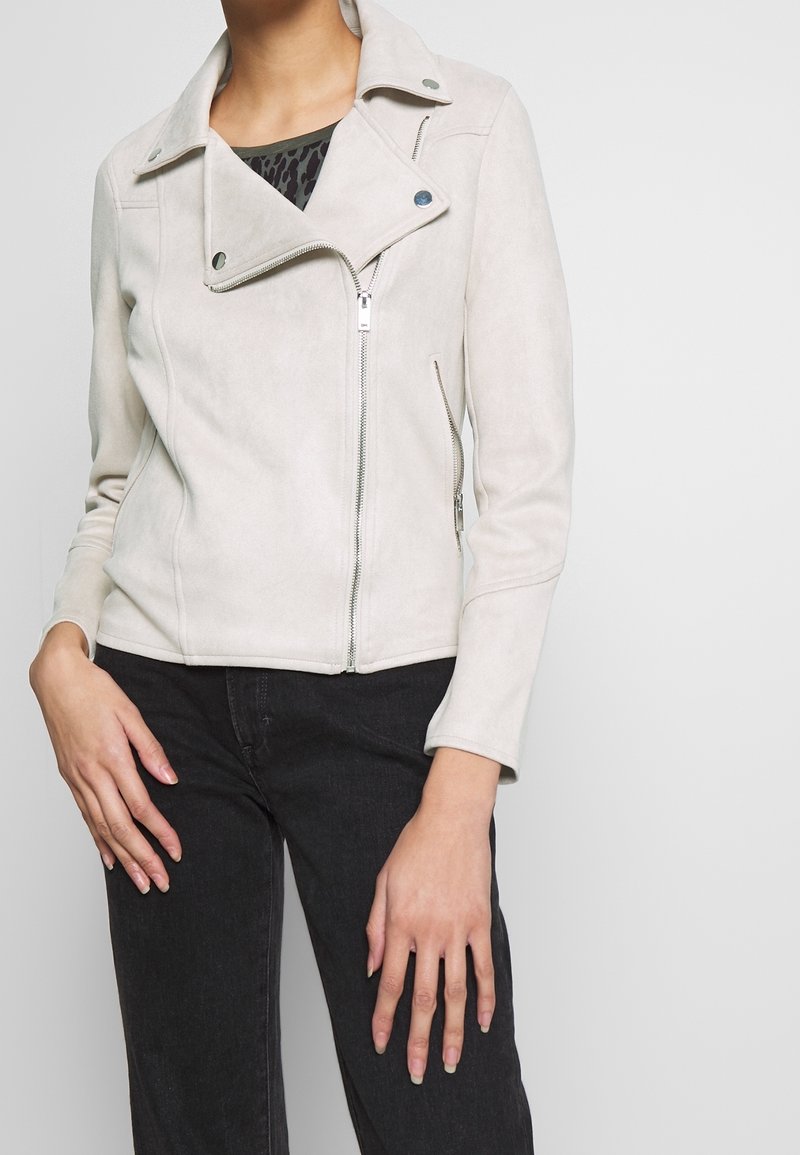 Motocyklowa kurtka z jasnoszarego sztucznego zamszu z zamkiem błyskawicznym, kołnierzem na zatrzask i skośnymi kieszeniami. Charakteryzuje się dopasowanym krojem i gładką fakturą.