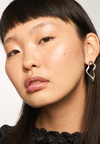 Boucles d'oreilles argentées en forme abstraite avec un design ondulé. Texture lisse sur un tissu sombre contrastant. Éclat sur la peau.