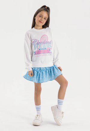 Weißes Sweatshirt mit buntem "Cleveland Tennis"-Design, kombiniert mit einem blauen, floral gemusterten Rüschenschein, weißen Sneakers und gestreiften Socken.