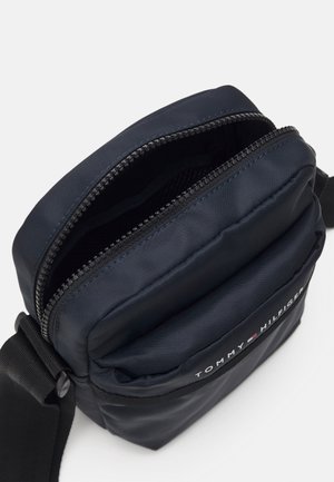 Lille navyblå crossbody taske med åben sort lynlås, forlommer og justerbar sort rem, mærket med Tommy Hilfiger-logo.