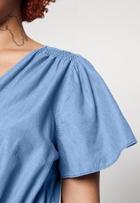 Femme portant un chemisier bleu clair à manches courtes avec des épaules froncées et un col en V, montrant un tatouage dans le haut du dos et le cou.