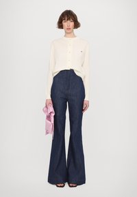 Krämfärgad kardigan med knäppning, högmidjade mörka denimbyxor med utsvängda ben och en ljusrosa handväska med texturerad yta.
