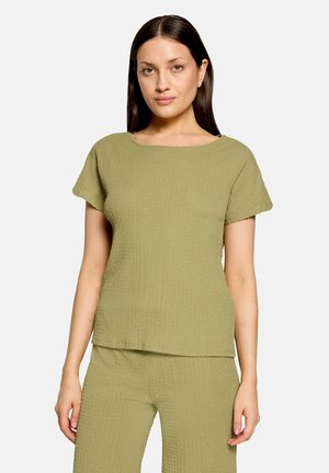 Femme aux longs cheveux foncés portant un haut à manches courtes texturé vert olive clair et un pantalon assorti, debout devant un fond uni.