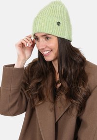 Groene geribbelde gebreide beanie met een opgevouwen boord en een klein zwart logo-detail, gedragen door een persoon met lang zwart haar, gecombineerd met een bruine jas.