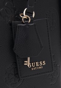 Sac en cuir noir texturé avec breloque clé dorée et étiquette de marque Guess embossée de "EST 1981" en lettres dorées.