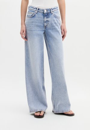 Femme portant un jean denim large de couleur bleu clair et des tongs noires, se tenant devant un fond blanc uni.