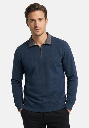 Mand iført en mørkeblå langærmet pullover med kvartlynlås og krave, stående med hænderne i lommerne på sorte bukser.