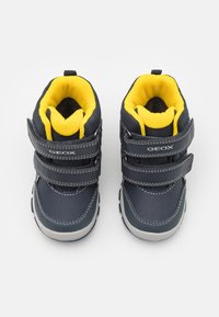 Zapatos infantiles de color azul oscuro con forro de malla amarilla, que cuentan con dos correas de Velcro ajustables y suelas de goma gris. Material superior texturizado.