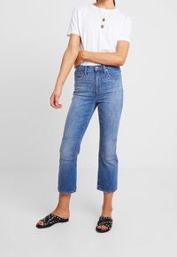 Ljust blå, högmidjade jeans med rak passform och knäpatchar. För att komplettera outfiten bärs de med en vit T-shirt och svarta slip-in sandaler med nitar.