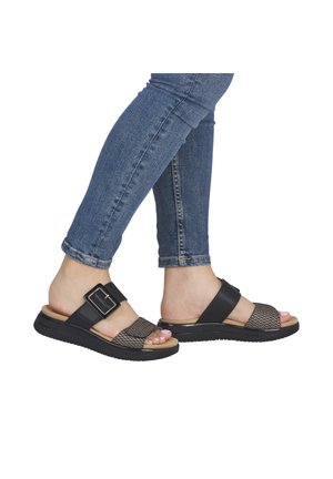 Feet wearing black open-toe sandals with buckle and woven straps, paired with blue skinny jeans against a white background.