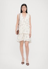 Ung kvinne iført en ermeløs hvit blomstret kjole med volanger, brune åpne pumps og bærende en brun skulderveske.