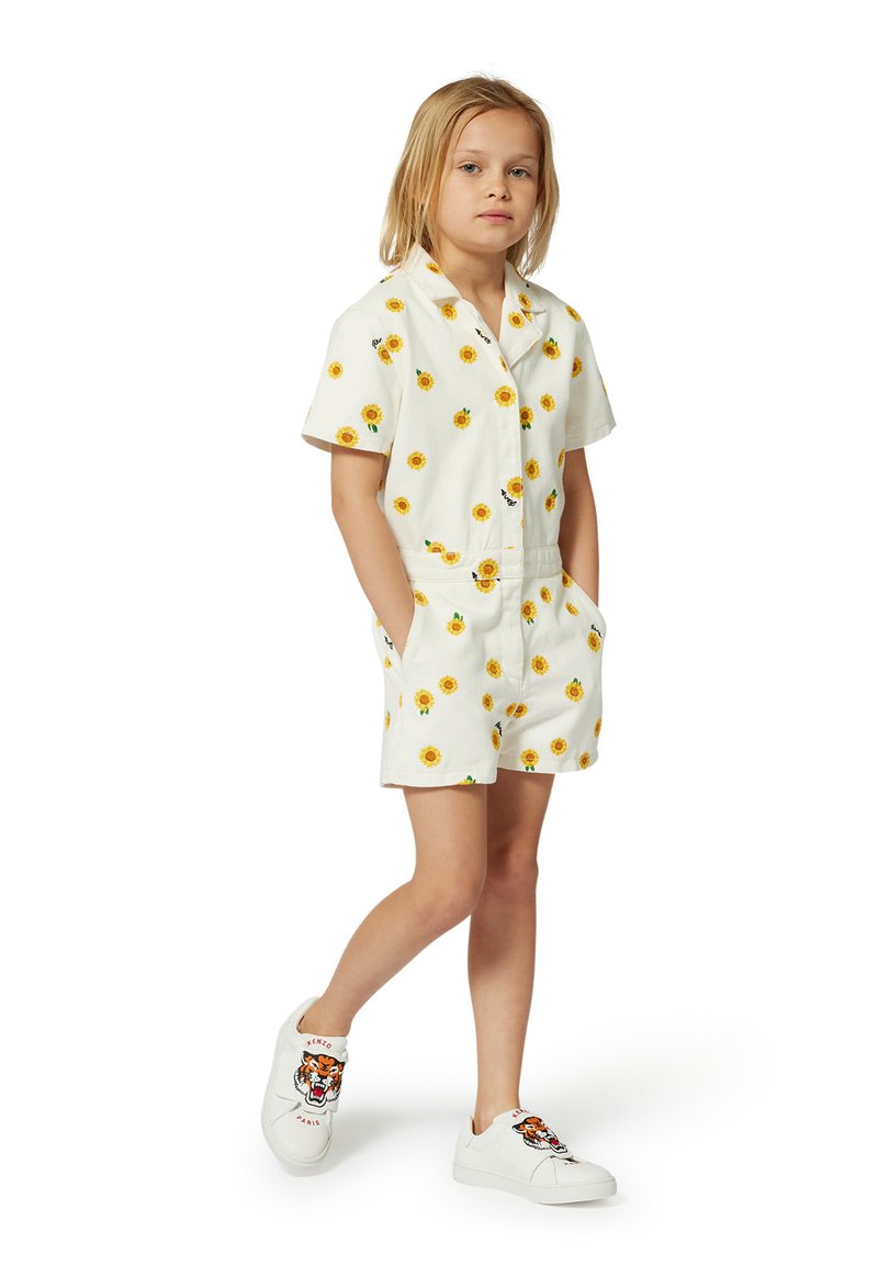 Combinaison courte blanche avec imprimé tournesol, manches courtes, boutonnière à l'avant, taille élastique et poches latérales. Associée à des chaussures blanches avec un design de tigre.