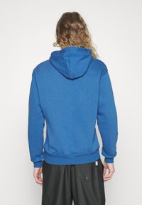 Osoba s dlouhými vlasy, oblečená v modré mikině s kapucí a černých kalhotách, stojící zády k fotoaparátu na jednolitém bílém pozadí.