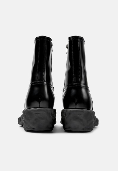 Botas negras de cuero hasta el tobillo con un acabado suave, cremalleras traseras y suelas negras gruesas y texturizadas. Detalle de costura simple a lo largo de las costuras.