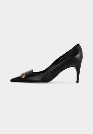 Schwarzer spitzer High-Heel-Schuh mit strukturiertem Muster und kleinem goldenen Schnallendetail vorne, seitlich auf einfarbigem Hintergrund dargestellt.
