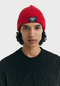 Bonnet rouge côtelé en tricot avec une étiquette portant l'inscription "THINKING MU". Le modèle porte un pull foncé et texturé. Le fond neutre met en valeur les accessoires.