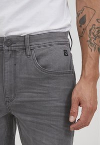 Vaqueros de denim gris con un diseño estilizado, cierre de botón y un pequeño parche con el logo en el bolsillo. La tela tiene una textura suave.