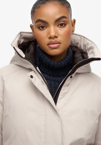 Beżowy, ocieplany płaszcz z wysokim kołnierzem i kapturem. Posiada zamek błyskawiczny oraz ciemnoniebieski, dziany golf pod spodem. Gładka, matowa faktura.