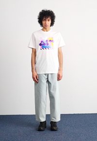 Λευκό βαμβακερό t-shirt με πολύχρωμο αφηρημένο γραφικό, συνδυασμένο με ανοιχτό μπλε φαρδύ παντελόνι και μαύρα αθλητικά παπούτσια, σε απλό φόντο.