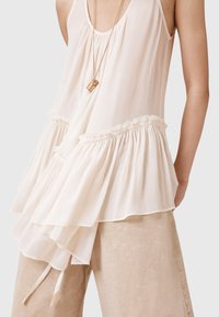 Crème-kleurig mouwloos topje met gelaagde ruffle en gestructureerde stof, gecombineerd met lichtbeige linnen broek. Lange, dunne bandjes hangen van de top.
