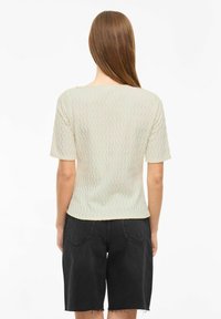 Top corto de manga corta en color beige claro con un patrón texturizado en ondas, con un escote redondeado, combinado con pantalones cortos de mezclilla gris oscuro.
