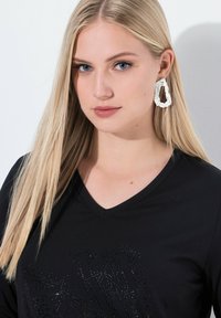 Černý top s výstřihem do V, který má texturovaný design s kamínky; modelka má na sobě velké bílé abstraktní náušnice; rovné blond vlasy.
