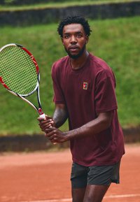 Tennispelaaja, joka pitää mailaa, päällään marooni T-paita, jossa on pieni logo, ja tummat shortsit, savikentällä vihreän taustan kanssa.