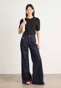 Μαύρη μπλούζα με δαντελένια φουσκωτά μανίκια, συνδυασμένη με ψηλόμεσα, φαρδιά τζην σε σκούρο χρώμα. Το μοντέλο φορά ροζ παπούτσια και κρατά μια ανοιχτόχρωμη τσάντα.