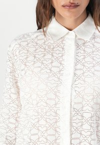 Lett hvit skjorte med et subtilt geometrisk mønster i beige. Har klassisk krage og knappelukking foran. Mykt, teksturert stoff.