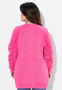Sudadera rosa con mangas texturizadas y fruncidas, y cuello acanalado, presenta un corte holgado y una longitud larga, fabricada con tela suave.