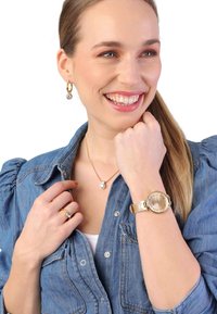 Gouden horloge met een gestructureerde wijzerplaat, lichtbeige band, zilveren ketting met een ronde hanger en bijpassende ring, gedragen bij een denim shirt.