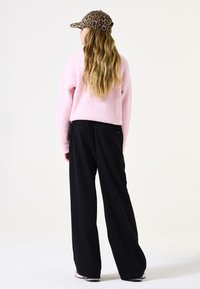 Roze geribde trui, zwarte wijde broek en een cap met luipaardprint, gezien vanuit de rug, waarbij de ontspannen pasvorm en textuurdetails zichtbaar zijn.