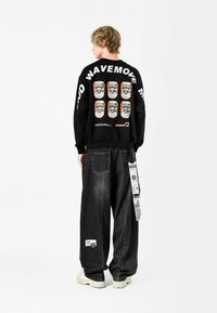 Sudadera negra con un diseño gráfico en la parte posterior que presenta múltiples ilustraciones de latas y texto, combinada con unos vaqueros grises de corte holgado.