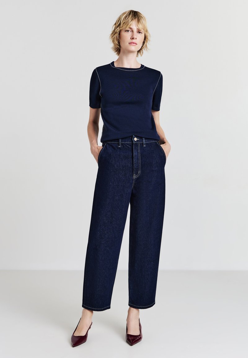 Donna bionda in piedi con le mani in tasca, che indossa una maglietta blu navy a maniche corte, jeans larghi scuri e décolleté borgogna su uno sfondo semplice.