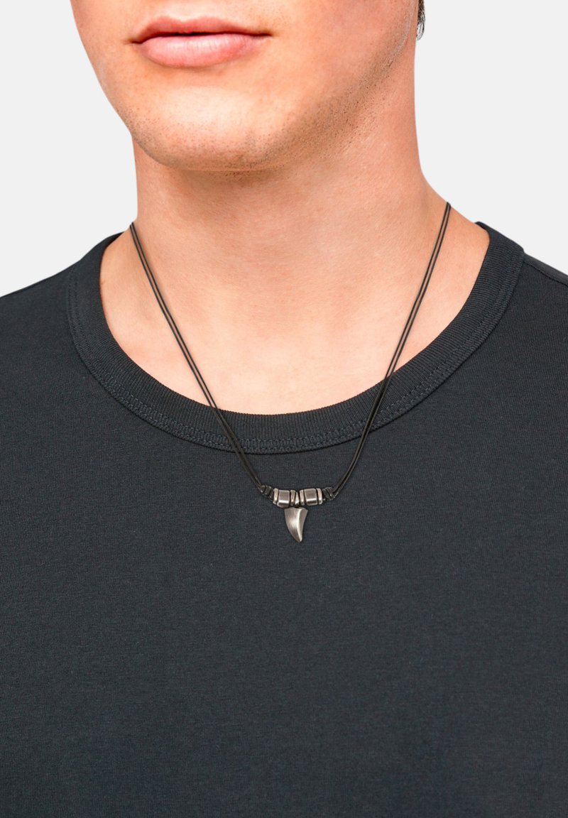 Collana nera con pendente a dente di squalo e piccole perline metalliche. Indossata sopra una maglia scura con scollo a giro. Texture liscia.