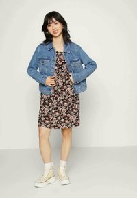 Jaqueta de ganga sobre um vestido floral, com padrões em rosa e branco. A modelo usa sapatilhas altas brancas, em pé contra um fundo claro.