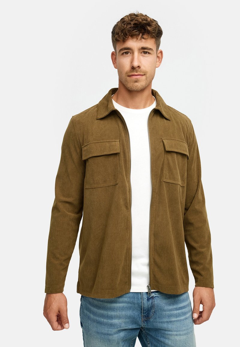Mand iført brun fløjlsjakke over hvid T-shirt og blå jeans, stående mod en ensfarvet hvid baggrund.