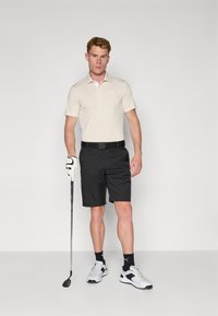 Beige Poloshirt mit kurzen Ärmeln, schwarze Shorts und weiße Golfhandschuhe. Stehend vor einem grauen Hintergrund, trägt sportliche Schuhe.