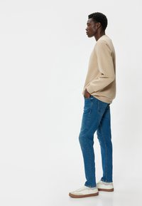 Sudadera beige de manga larga, jeans azul ajustados y zapatillas blancas con detalles marrones, mostradas en un modelo de perfil.