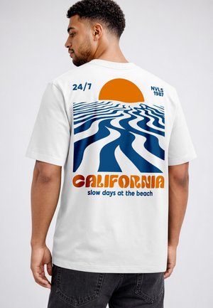 Man wearing white t-shirt with orange sun and blue abstract waves, text "California slow days at the beach," and "24/7 NVLS 1987."