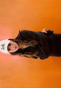 Schwarze Zip-up-Sweatjacke, taillierte Silhouette, über einem schwarzen Rock. Weiße Kappe mit gesticktem "LA", runde Ohrringe, strukturierter oranger Hintergrund.
