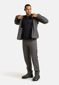 Grå letvægtsjakke med lynlås fortil, parret med en sort T-shirt med et blåt campingmotiv, og grå bukser med lynlås lommer.