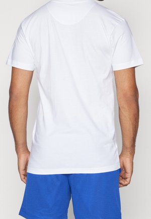 Mężczyzna w prostym białym t-shircie z krótkim rękawem i niebieskich sportowych szortach z siateczki, pokazany od tyłu na jednolitym tle.
