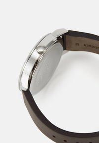 Reloj de tono plateado con caja redonda, correa de cuero marrón oscuro y corona de acero inoxidable, que presenta un acabado suave y un diseño minimalista.