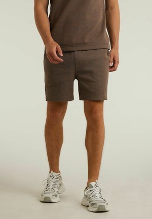 Man draagt bruine shorts en bijpassend shirt met witte sportsneakers en staat op een lichte achtergrond.
