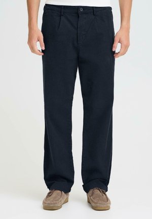 Mees, kes kannab lahtisi musti pükse ja pruune seemisnahast mokasiinsussisid, seisab tasasel heletaustal.