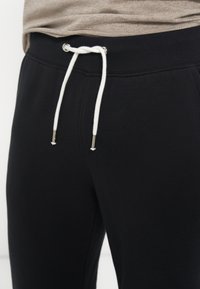 Pantalon de survêtement noir avec une taille élastique côtelée et un cordon blanc noué en un nœud lâche, porté avec un haut beige.