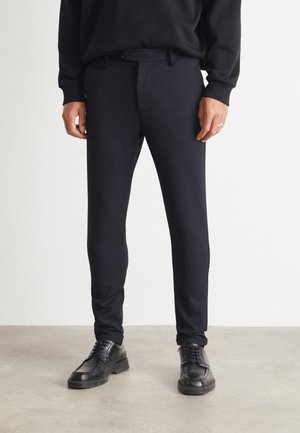 Homme portant un pantalon noir slim et des chaussures en cuir noir à lacets, debout sur un sol gris contre un mur blanc.