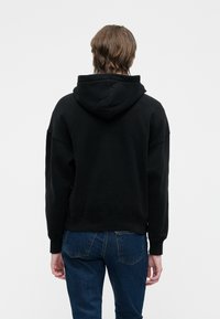 Lühikeste pruunide juustega inimene, kes kannab musta kapuutsi ja siniseid teksapükse, seisab seljaga kaamera poole tavalise valge tausta vastu.