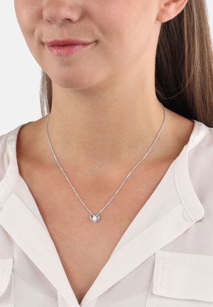 Collana d'argento a forma di cuore su una delicata catena, con un piccolo dettaglio intagliato. Indossata su una blusa di colore chiaro.